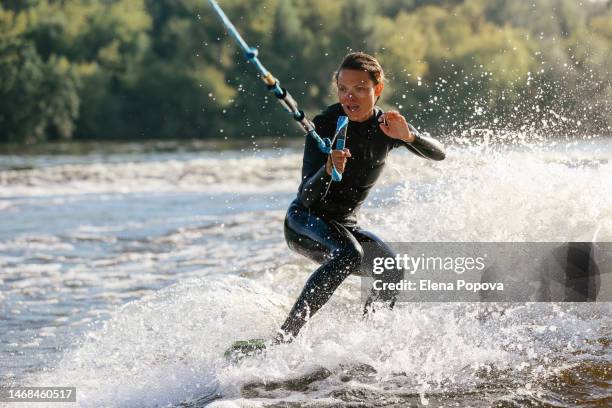 mid adult female surfer wakeboarding against blurred riverside background on sunny summer day - wakeboarden stock-fotos und bilder