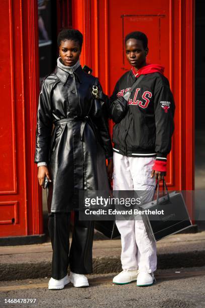 Model wears a black shiny leather high belted long coat, a pale gray ribbed wool pullover, black shiny leather flared pants, white leather sneakers,...