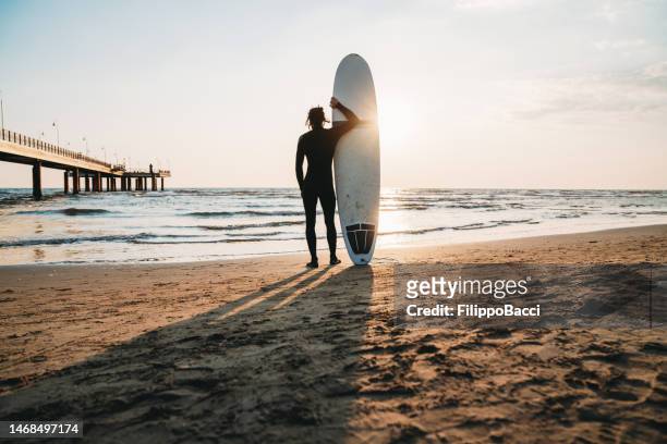 ein mann steht mit seinem surfbrett am strand und bewundert den sonnenuntergang - beach hold surfboard stock-fotos und bilder