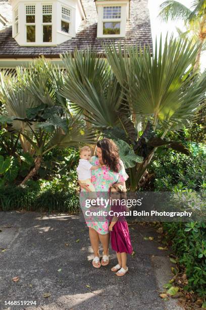 a happy 31-year-old cuban mother with curly brown hair & wearing a colorful spring dress while holding her snuggly baby boy & standing by her 3-year-old daughter while walking peacefully on a lake trail in palm beach, fl - snuggly stock pictures, royalty-free photos & images