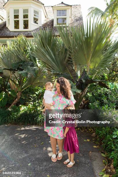 a happy 31-year-old cuban mother with curly brown hair & wearing a colorful spring dress while holding her snuggly baby boy & standing by her 3-year-old daughter while walking peacefully on a lake trail in palm beach, fl - snuggly stockfoto's en -beelden