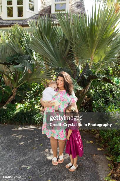 a happy 31-year-old cuban mother with curly brown hair & wearing a colorful spring dress while holding her snuggly baby boy & standing by her 3-year-old daughter while walking peacefully on a lake trail in palm beach, fl - snuggly stock pictures, royalty-free photos & images