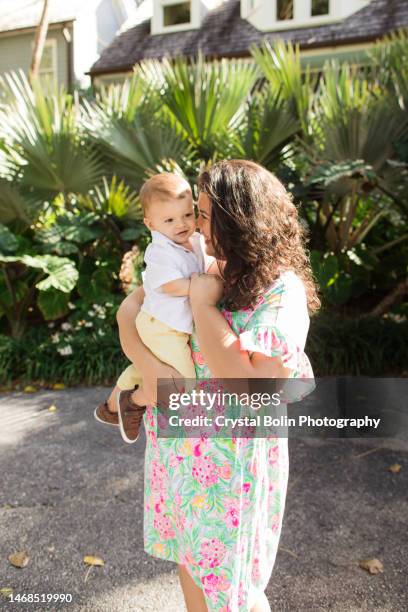 a happy 31-year-old cuban mother with curly brown hair & wearing a colorful spring dress while holding her snuggly baby boy dressed in a white short sleeve shirt, light yellow pants & brown shoes while walking peacefully on a lake trail in palm beach, fl - snuggly stock pictures, royalty-free photos & images