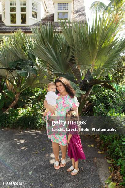 a happy 31-year-old cuban mother with curly brown hair & wearing a colorful spring dress while holding her snuggly baby boy & standing by her 3-year-old daughter while walking peacefully on a lake trail in palm beach, fl - snuggly stockfoto's en -beelden