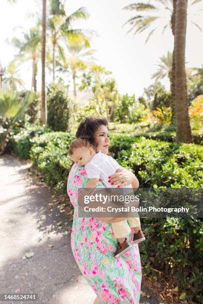 a happy 31-year-old cuban mother with curly brown hair & wearing a colorful spring dress while holding her snuggly baby boy dressed in a white short sleeve shirt, light yellow pants & brown shoes while walking peacefully on a lake trail in palm beach, fl - snuggly stock pictures, royalty-free photos & images