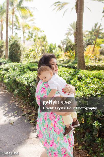 a happy 31-year-old cuban mother with curly brown hair & wearing a colorful spring dress while holding her snuggly baby boy dressed in a white short sleeve shirt, light yellow pants & brown shoes while walking peacefully on a lake trail in palm beach, fl - snuggly stock pictures, royalty-free photos & images