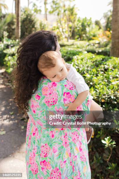 a happy 31-year-old cuban mother with curly brown hair & wearing a colorful spring dress while holding her snuggly baby boy dressed in a white short sleeve shirt, light yellow pants & brown shoes while walking peacefully on a lake trail in palm beach, fl - snuggly stock pictures, royalty-free photos & images