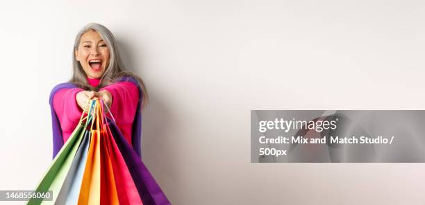 excited korean lady with grey hair,laughing and showing shopping bags - lady grey background bildbanksfoton och bilder