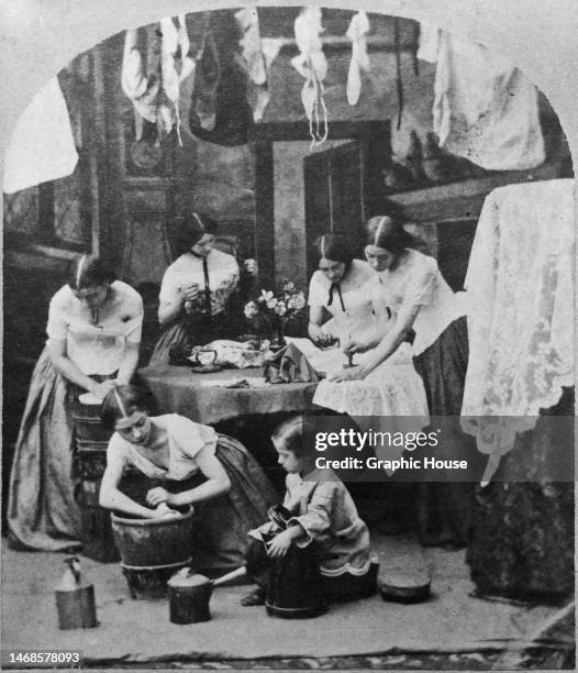 Group of women and girls at work in a laundry, with two women to the right of the frame ironing on a table, with wet laundry hanging to dry from the...
