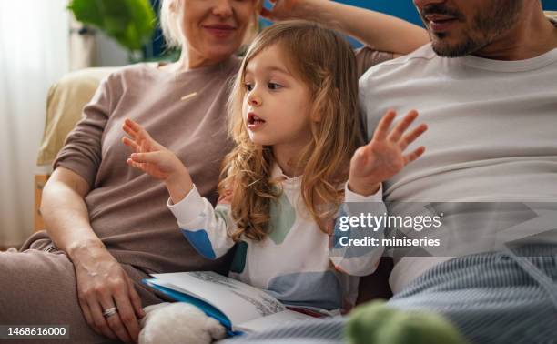 happy little girl reading a book with her parents while sitting in the bedroom together - 3 year old stock pictures, royalty-free photos & images