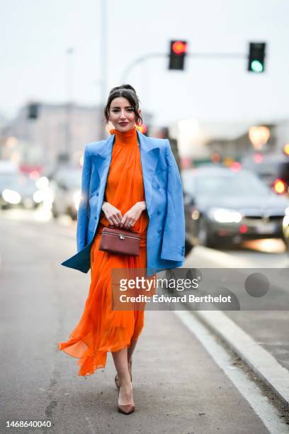 Alexandra Pereira wears a neon orange halter high neck / backless / long silk dress, a blue blazer jacket, diamonds rings, a brown shiny leathers...