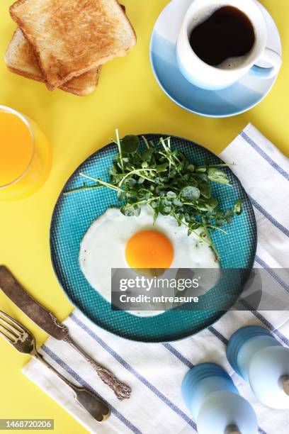 bild eines türkisfarbenen tellers mit sonnigem spiegelei mit brunnenkresse, kaffeetasse und untertasse, glas orangensaft, zwei scheiben weißem toast, messer und gabel, geschirrtuch, salz- und pfefferstreuer, gelber hintergrund, erhöhte aussicht - orange juice glass white background stock-fotos und bilder