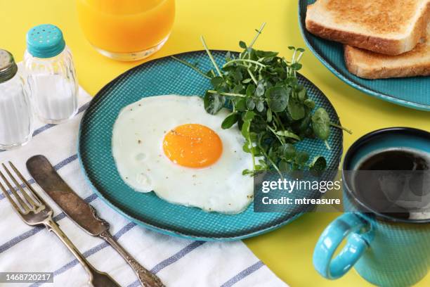 bild eines türkisfarbenen tellers mit sonnigem spiegelei mit brunnenkresse, tasse schwarzem kaffee, glas orangensaft, zwei scheiben weißem toast, messer und gabel, geschirrtuch, salz- und pfefferstreuer, gelber hintergrund, fokus auf vordergrund - orange juice glass white background stock-fotos und bilder