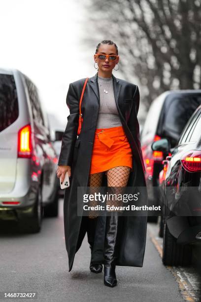 Guest wears neon orange sunglasses, gold necklaces, a pale gray high neck pullover, a black shiny leather long coat, a neon orange short skirt, black...