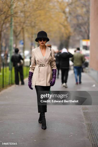 Guest wears black felt wool hat, black sunglasses, a beige belted jacket from Fendi, dark purple shiny leather gloves, a black tube midi skirt, black...