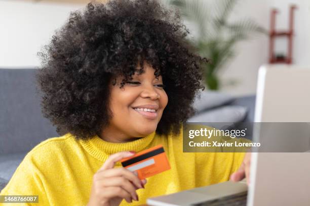 beautiful young latin american woman with afro hairstyle and yellow shirt holding credit card while using laptop computer for online shopping. - chubby credit fotografías e imágenes de stock