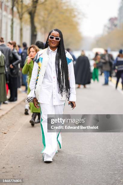 Guest wears beige plastic sunglasses, silver earrings, a white shirt with embroidered blue and red flower pattern details, a white latte with yellow...