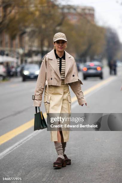 Guest wears a beige and dark brown monogram print pattern cap, yellow leather and gold earrings, a beige ribbed wool and black buttoned cardigan, a...