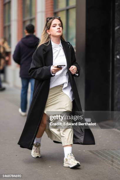 Guest wears black sunglasses, gold earrings, a white shirt, a black wool long coat, a black shiny leather shoulder bag, beige silk slit / split long...