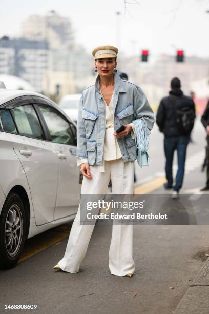 Guest wears a beige cap, gray silver pendant lion earrings, a blue faded denim cargo jacket, a white latte V-neck / silk belted jacket, matching...