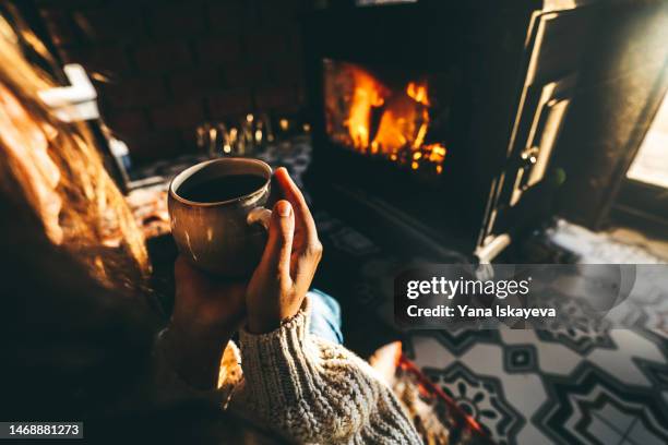 woman relaxing in front of the fireplace, embracing a mug with a hot drink - foyer de cheminée photos et images de collection