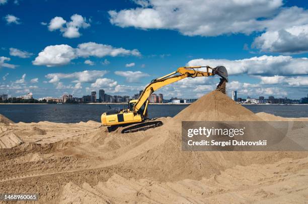 excavator on the bank of the river in the city digs the sand of the beach - sand pile stock pictures, royalty-free photos & images