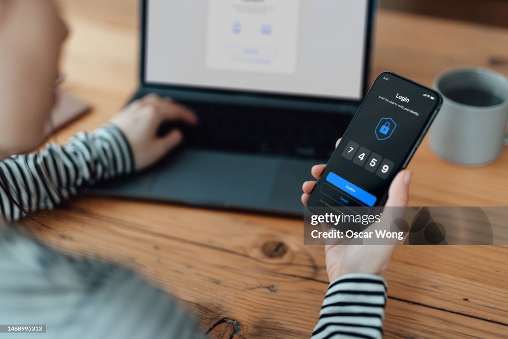 Over the shoulder view of young woman using mobile app with Two-Factor Authentication (2FA) security system to access online bank account via laptop