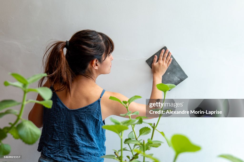 Woman Repairs White Wall Using Sandpaper near Plants