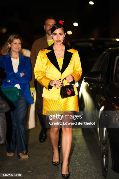 Bettina Looney wears gold earrings, a gold chain with a crystal pendant necklace, a yellow shoulder-off silk short tube dress, a matching yellow silk...