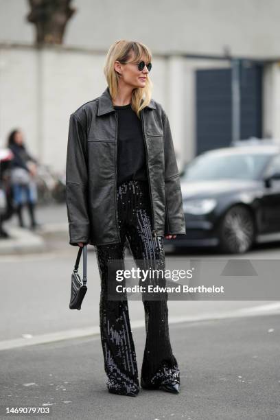 Jeanette Madsen wears black sunglasses, a black t-shirt, a black shiny leather oversized jacket, black embroidered sequined flared pants, black shiny...