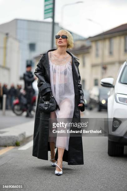 Viktoria Rader wears black sunglasses, silver triangle pendant earrings from Prada, a black shiny leather long coat, a black shiny leather handbag, a...