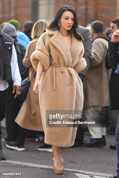 Jessica Wang wears gold earrings, gold necklaces, a beige sheep fur oversized long puffy coat, a beige shiny leather belt, beige shiny leather...