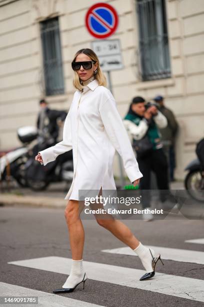 Candela Novembre wears black futurist sunglasses, silver earrings, a white shirt dress, a white latte turtleneck pullover, white socks, black shiny...