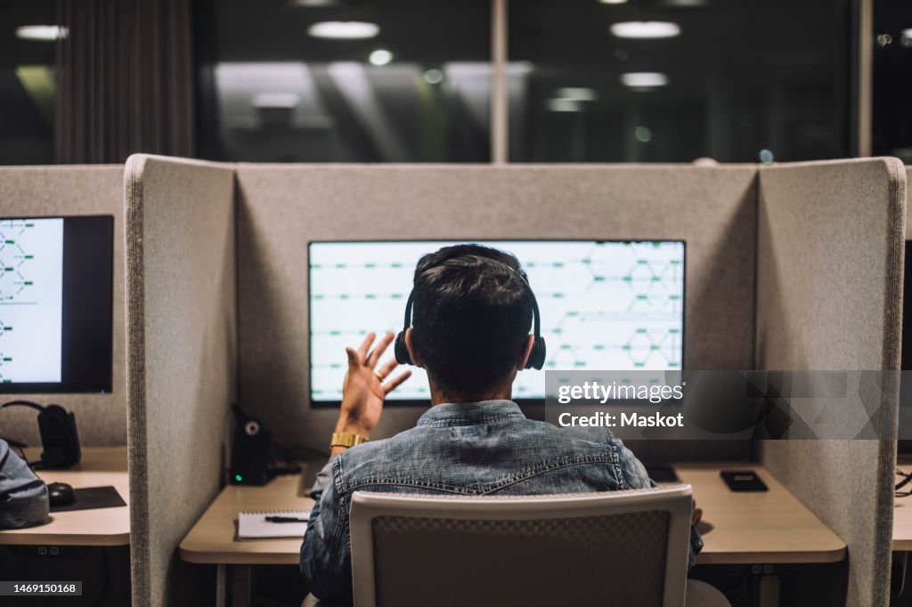 Rear view of young male customer service executive talking through headset sitting at desk in illuminated call center