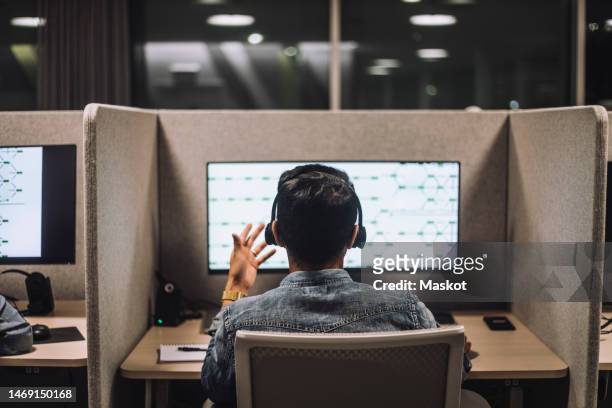rear view of young male customer service executive talking through headset sitting at desk in illuminated call center - call centre stock pictures, royalty-free photos & images