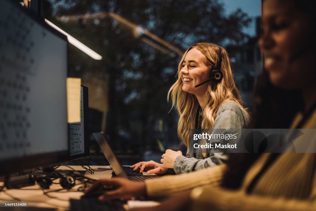 Cheerful young female customer service representative working by colleague at desk in call center