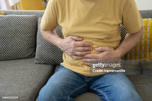 senior man with stomach pain sitting on couch in the living room - men stomach pain stock-fotos und bilder