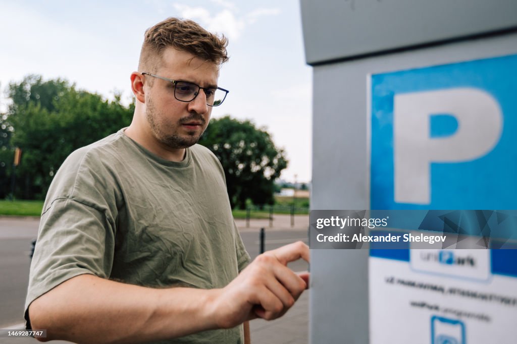 Person is using parking terminal for paying the station. Male portrait of man wear t-shirt and eyeglasses outdoors. Lifestyle photography concept.