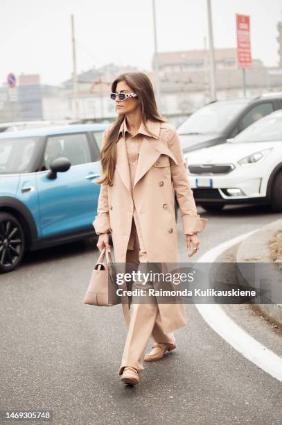 Clara Berry is seen wearing black and white sunglasses, a black and silver ring, gold rings, a light-beige monochrome look made of a long trench, a...