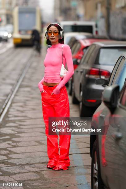 Alexandra Guerain wears silver headphones, a pale pink long sleeves t-shirt, a white shiny leather shoulder bag, orange sunglasses, red neon shiny...