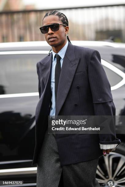Asap Rocky wears black sunglasses, a white shirt, a black tie, a black striped print pattern oversized blazer jacket, a gray slit / split long skirt,...