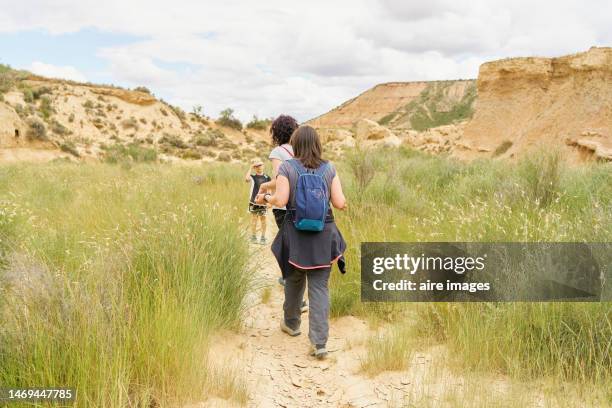 rear view of two women in casual clothes, one with a backpack and a child, walking along a path surrounded by vegetation and plants in bardenas reales rear view - comunidad foral de navarra fotografías e imágenes de stock