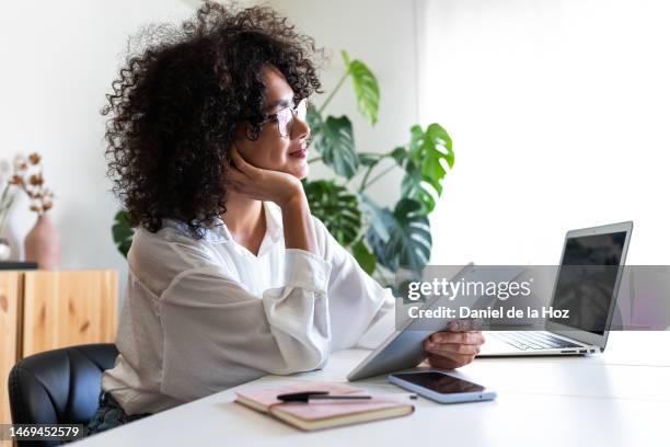 pensive, happy multiracial young woman working at home office using multiple devices: digital tablet, laptop and phone. - business woman blouse stock-fotos und bilder