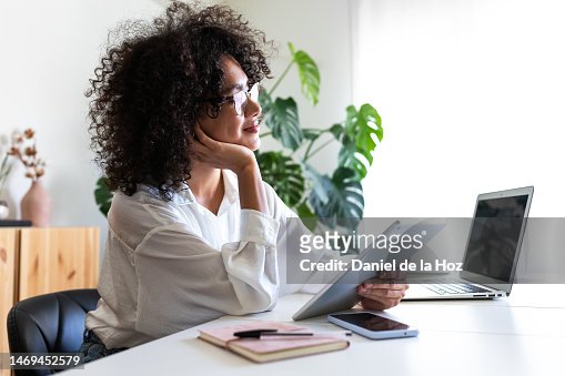 Pensive, happy multiracial young woman working at home office using multiple devices: digital tablet, laptop and phone.