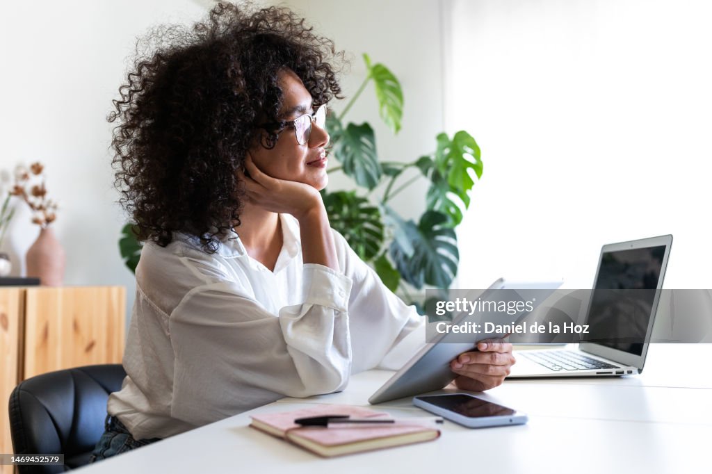 Pensive, happy multiracial young woman working at home office using multiple devices: digital tablet, laptop and phone.