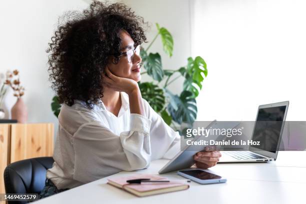 pensive, happy multiracial young woman working at home office using multiple devices: digital tablet, laptop and phone. - contemplation office stock pictures, royalty-free photos & images