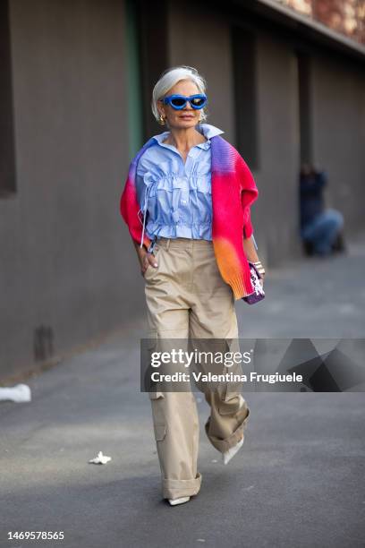 Grece Ghanem is seen wearing bright blue sunglasses, gold earrings, a cropped light blue shirt, a multicolored red, purple and orange sweater, cargo...