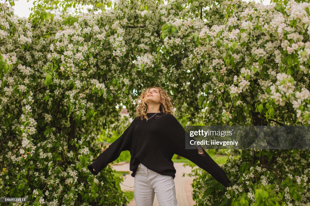 Blondes lockiges Mädchen in schwarzem Pullover auf dem Hintergrund eines blühenden Baumes. Garten. Weiße Blüten