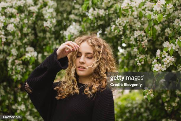 ragazza riccia bionda in maglione nero sullo sfondo dell'albero in fiore. giardino. fiori bianchi - closeup of a hispanic woman sneezing foto e immagini stock