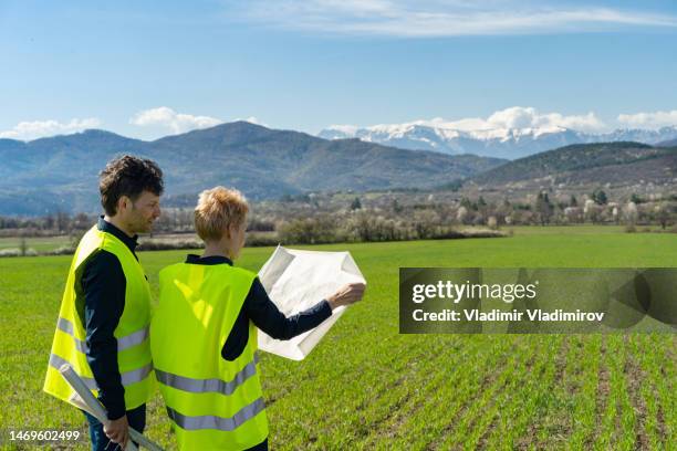 a man and a woman looking at blueprints standing in a field against mountains in the background - maatschappelijke verantwoordelijkheid stockfoto's en -beelden